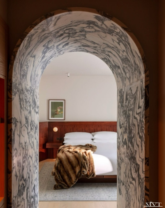 Gray-veined marble frames an arched entrance to a warm bedroom with white bedding, wood paneling, and a fur throw.