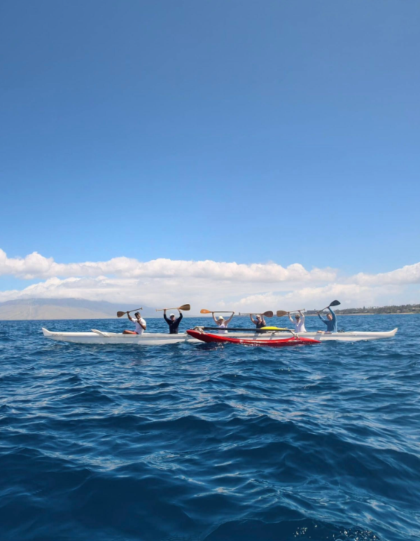 A group of paddlers raises oars from white canoes and a red kayak on deep blue water, with mountains in the distance.