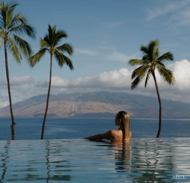 Infinity pool overlooks palm-lined coastline and mountains as a person rests in the water, facing the scenic ocean view.