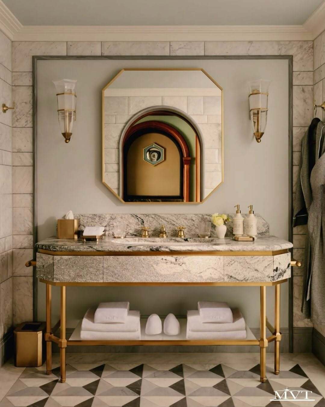Elegant marble vanity with gold accents, twin sconces, a large framed mirror, stacked towels, and patterned flooring.