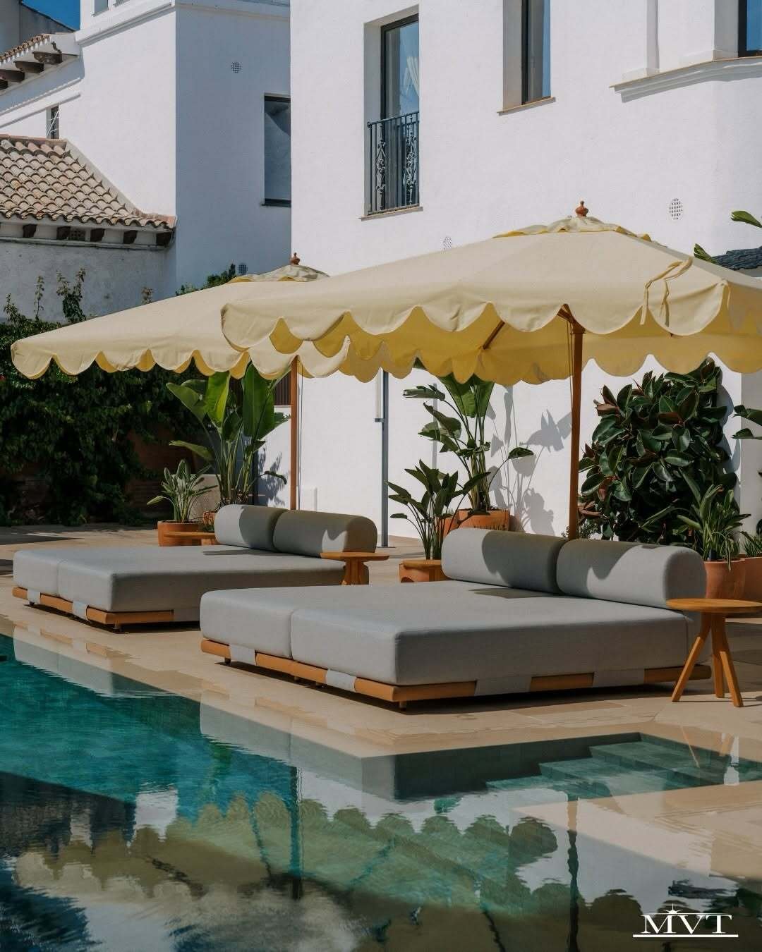 Two cushioned daybeds rest under scalloped cream umbrellas beside a turquoise pool and potted tropical greenery.