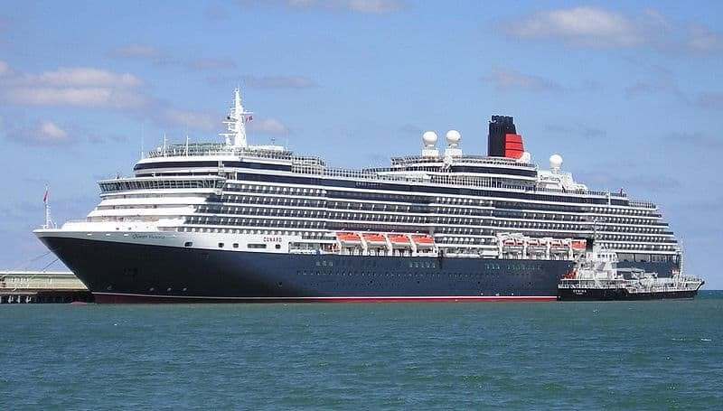 Large black and white cruise ship docked near a pier, featuring multiple passenger decks and orange lifeboats.