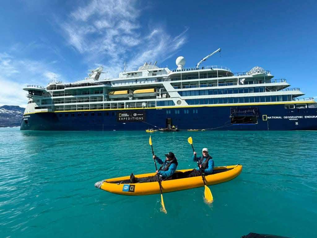 Two people paddle a yellow inflatable kayak through turquoise water beside a large National Geographic expedition ship.