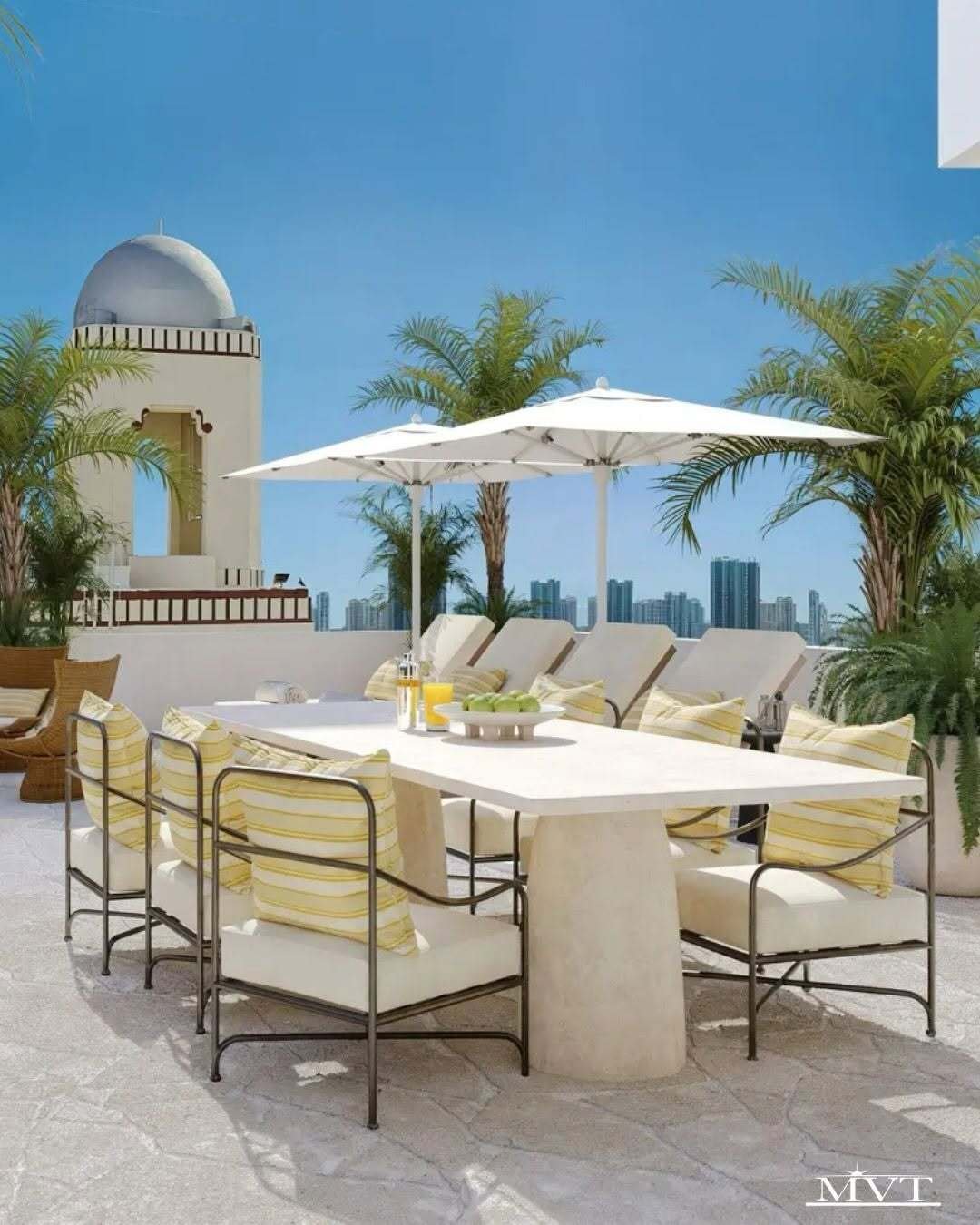 Luxury rooftop terrace with white dining furniture, striped cushions, palm trees, and umbrellas overlooking a city skyline.