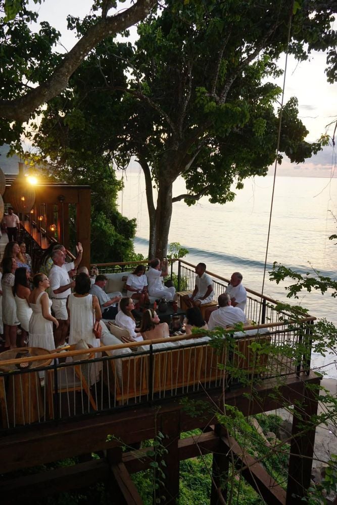 A group of guests socialize on an elevated seaside deck with cushioned seating, surrounded by trees and ocean views.