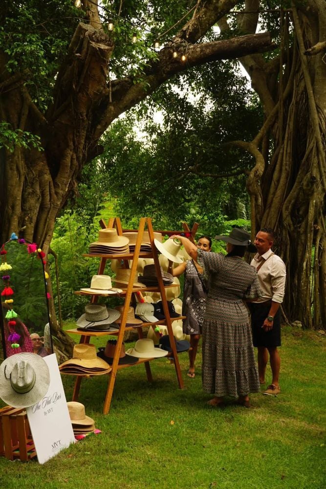 Guests browse a wooden display of assorted hats beneath towering trees, with colorful decorations hanging nearby.
