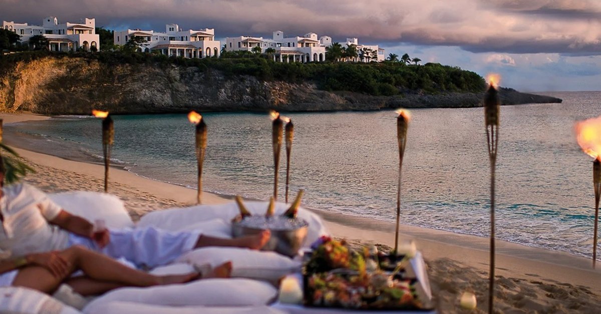 A couple relaxing on a private beachside lounge setup with ocean views, tiki torches, and luxury villas in the distance.