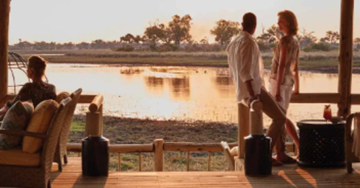People standing with luggage on a waterfront deck near chairs, calm water, trees, and warm sunlight.