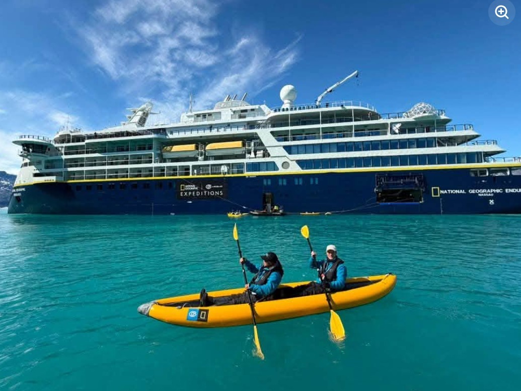 Two people paddle a yellow kayak on turquoise water beside the blue-and-white National Geographic Endurance ship.