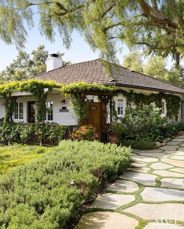 White cottage with a tiled roof and vine-covered porch, surrounded by lush gardens and a winding stone path.