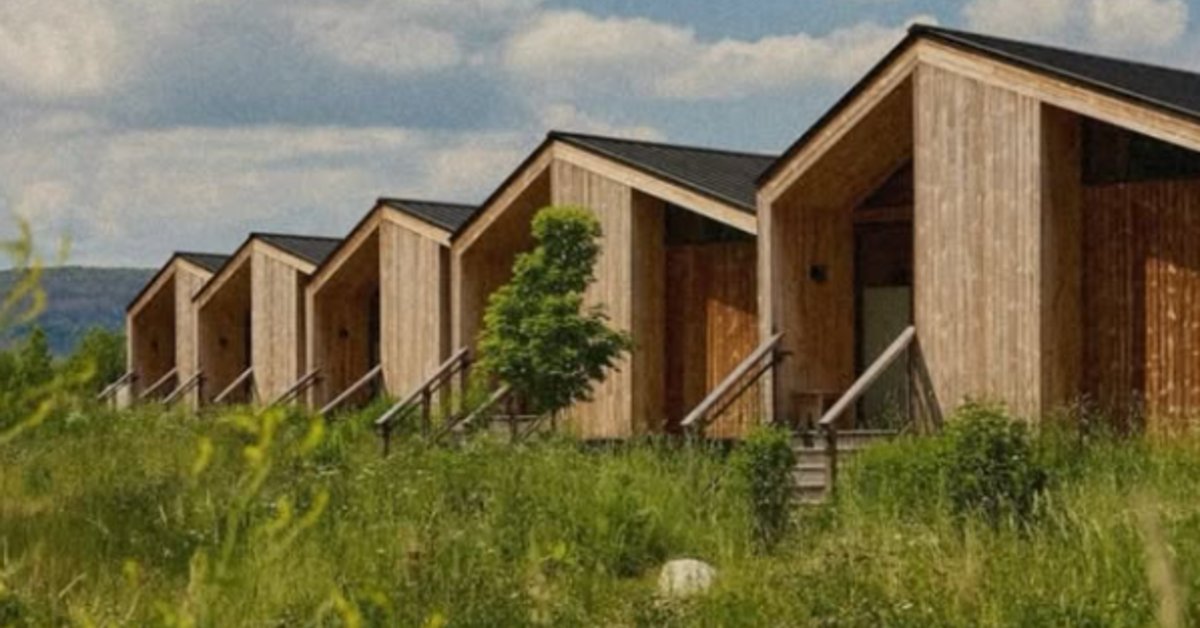 A row of wood cabins with sloped roofs beside tall grass, shrubs, mountains, and a cloudy sky behind them.