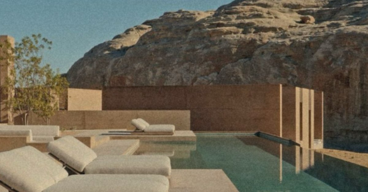 A modern outdoor pool area with cushioned lounge chairs, tan walls, rocky hills, and clear water under a blue sky.