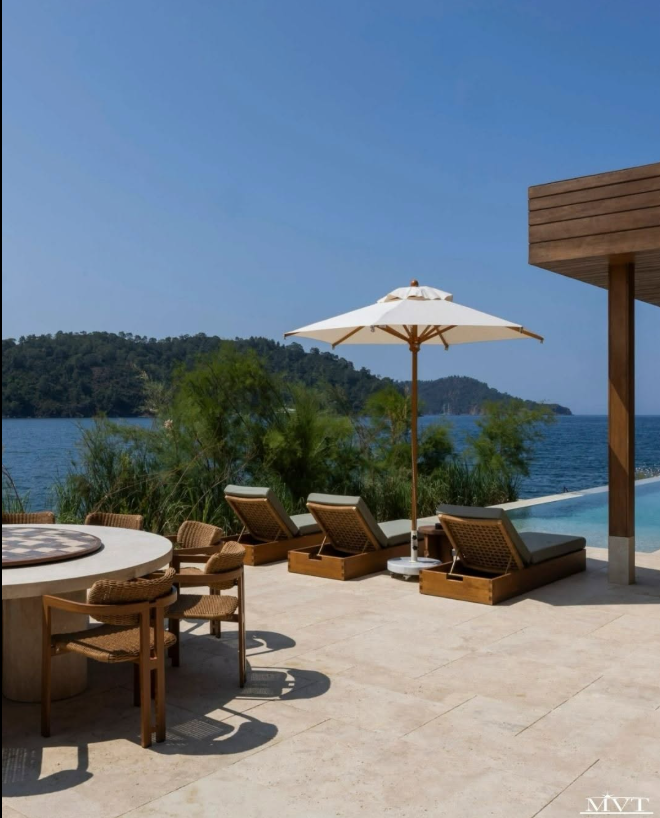Outdoor resort patio with cushioned loungers, a white umbrella, round dining table, blue sea, and wooded hills.