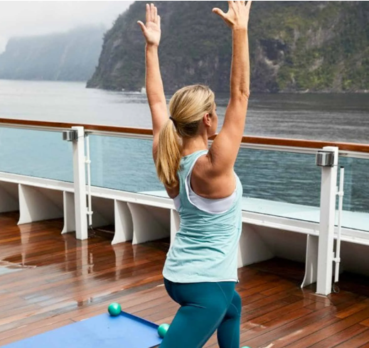 Woman stretches with arms raised on a wet wooden deck beside a yoga mat, overlooking calm water and cliffs.