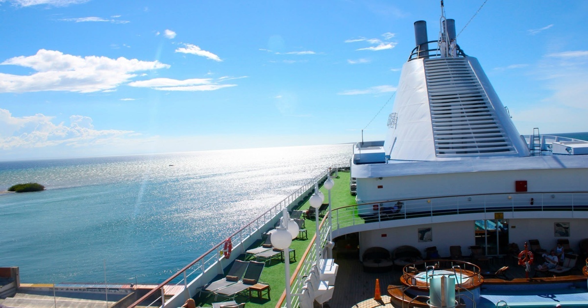 Upper deck of a cruise ship overlooks bright blue water, with pool seating, railings, and a white funnel.