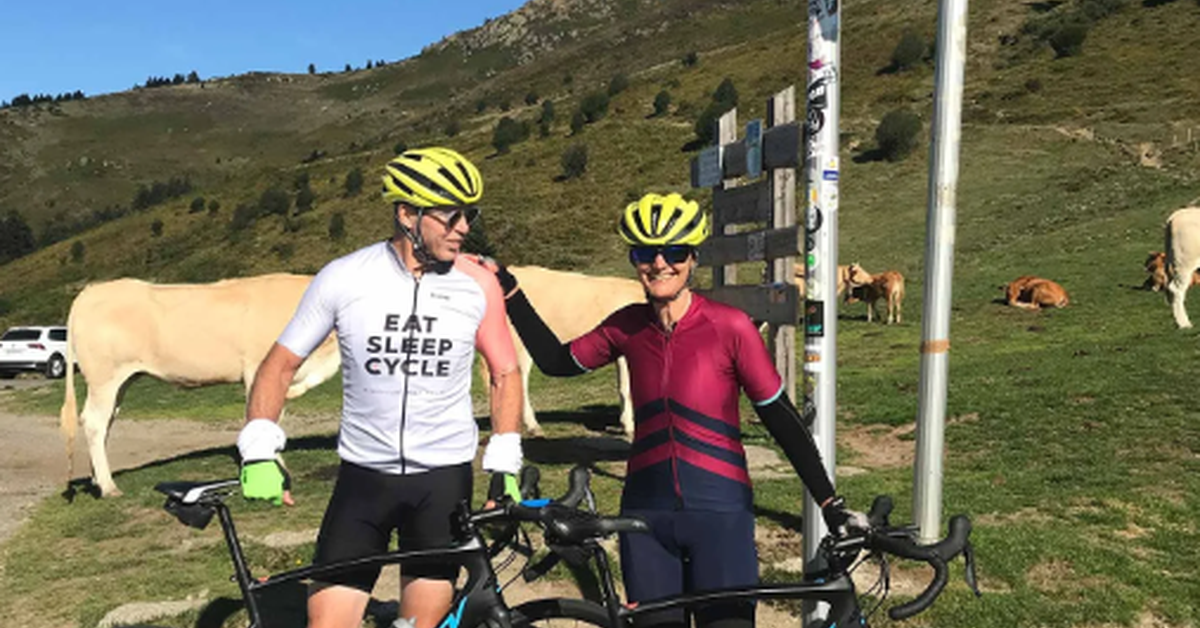 Two cyclists in helmets pose with road bikes beneath a mountain pass sign, with cows grazing on nearby hills.